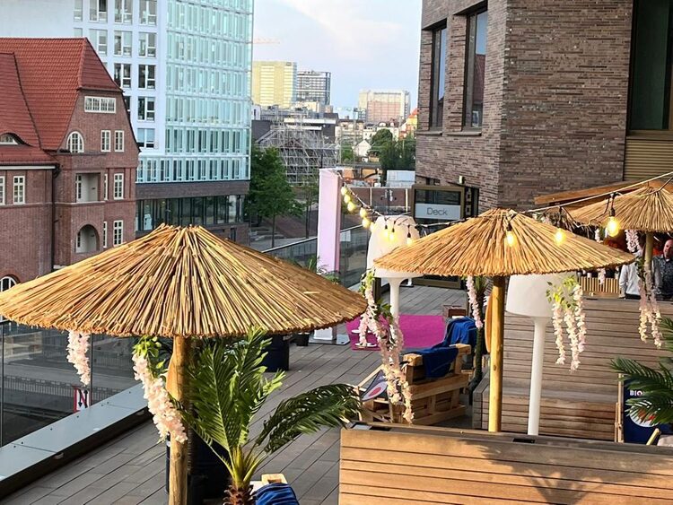 Dachterrasse mit strohgedeckten Sonnenschirmen, Palmen und Lichterkette, dahinter die Stadt