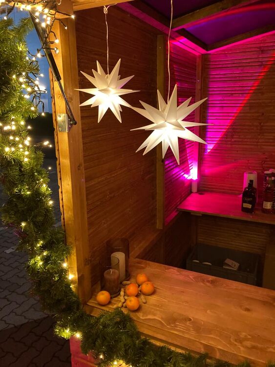 Zwei weiße Herrnhuter Sterne unter dem Hüttendach, dahinter magenta beleuchtete Holzwand, rechts die Theke mit Flaschen