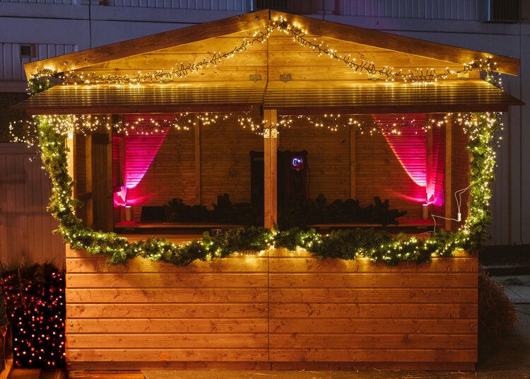Holzbude von vorn, zwei Verkaufsfenster magenta ausgeleuchtet, Girlande und Lichterketten an Giebel und Theke, links ein Tannenbaum